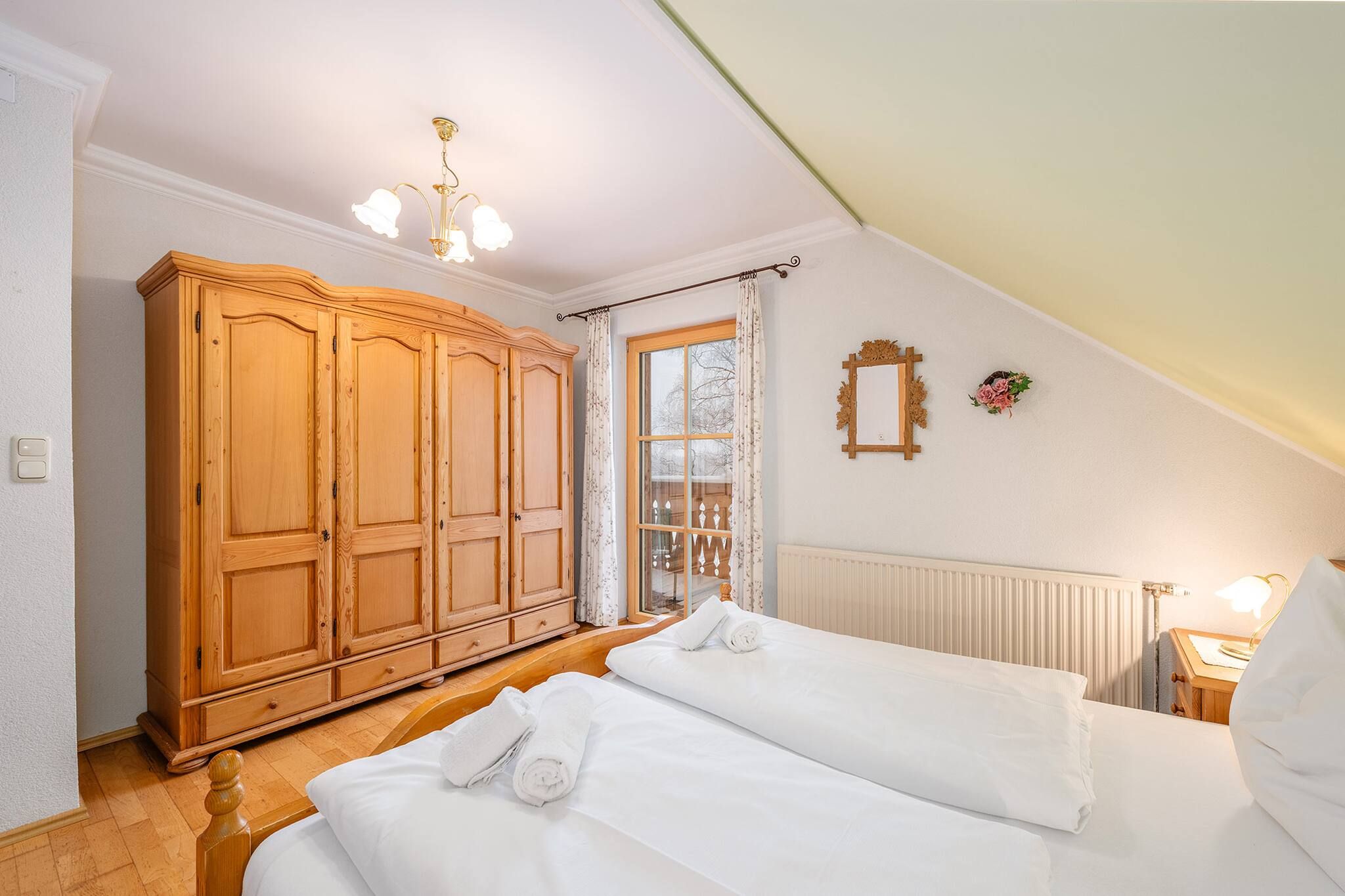 A bedroom with a large wardrobe, a bed with white linens, a window with curtains, and a wall-mounted mirror.