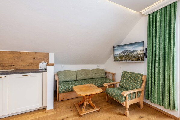 Ein kleiner, gemütlicher Raum mit Holzboden, einem grünen Sofa, einem Holzstuhl, einem kleinen Tisch und einer Küchenzeile mit einem Toaster.