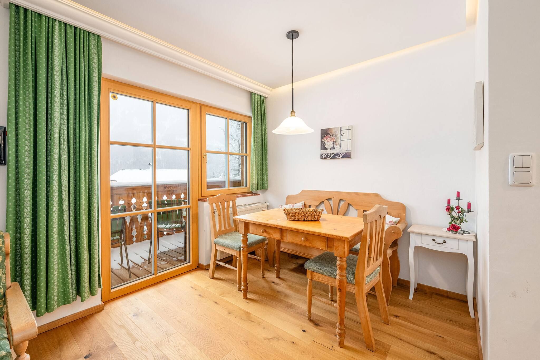 Ein Zimmer mit einem Holztisch, Stühlen, einer Couch, einem kleinen weißen Schreibtisch und einem Fenster mit grünen Vorhängen.