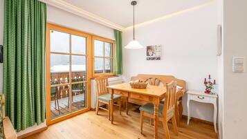 Ein Zimmer mit einem Holztisch, Stühlen, einer Couch, einem kleinen weißen Schreibtisch und einem Fenster mit grünen Vorhängen.