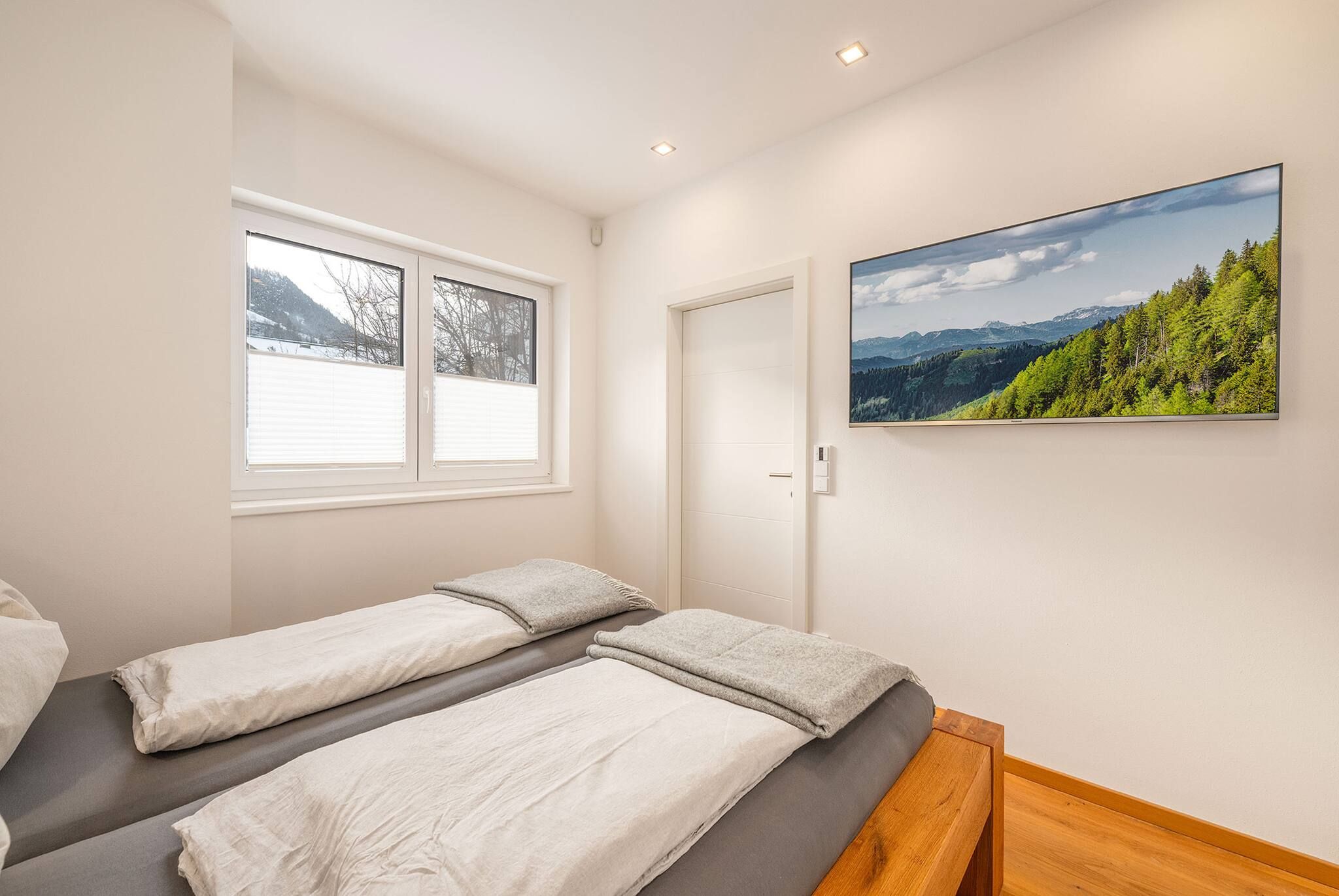 Un dormitorio con dos camas, una ventana con persianas, una puerta y un televisor de pared que muestra un paisaje.