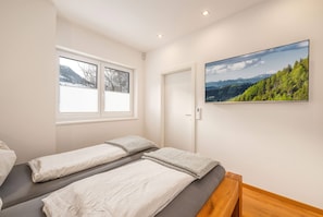 Un dormitorio con dos camas, una ventana con persianas, una puerta y un televisor de pared que muestra un paisaje.