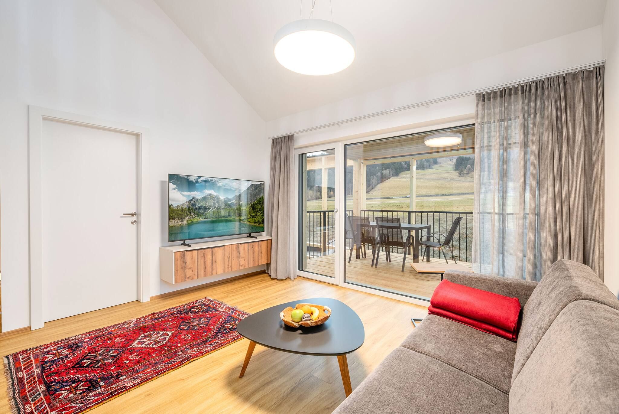Ein Wohnzimmer mit einer Couch, einem Couchtisch mit Obst, einem Fernseher auf einer Holzkommode und Blick auf einen Balkon mit Gartenmöbeln.