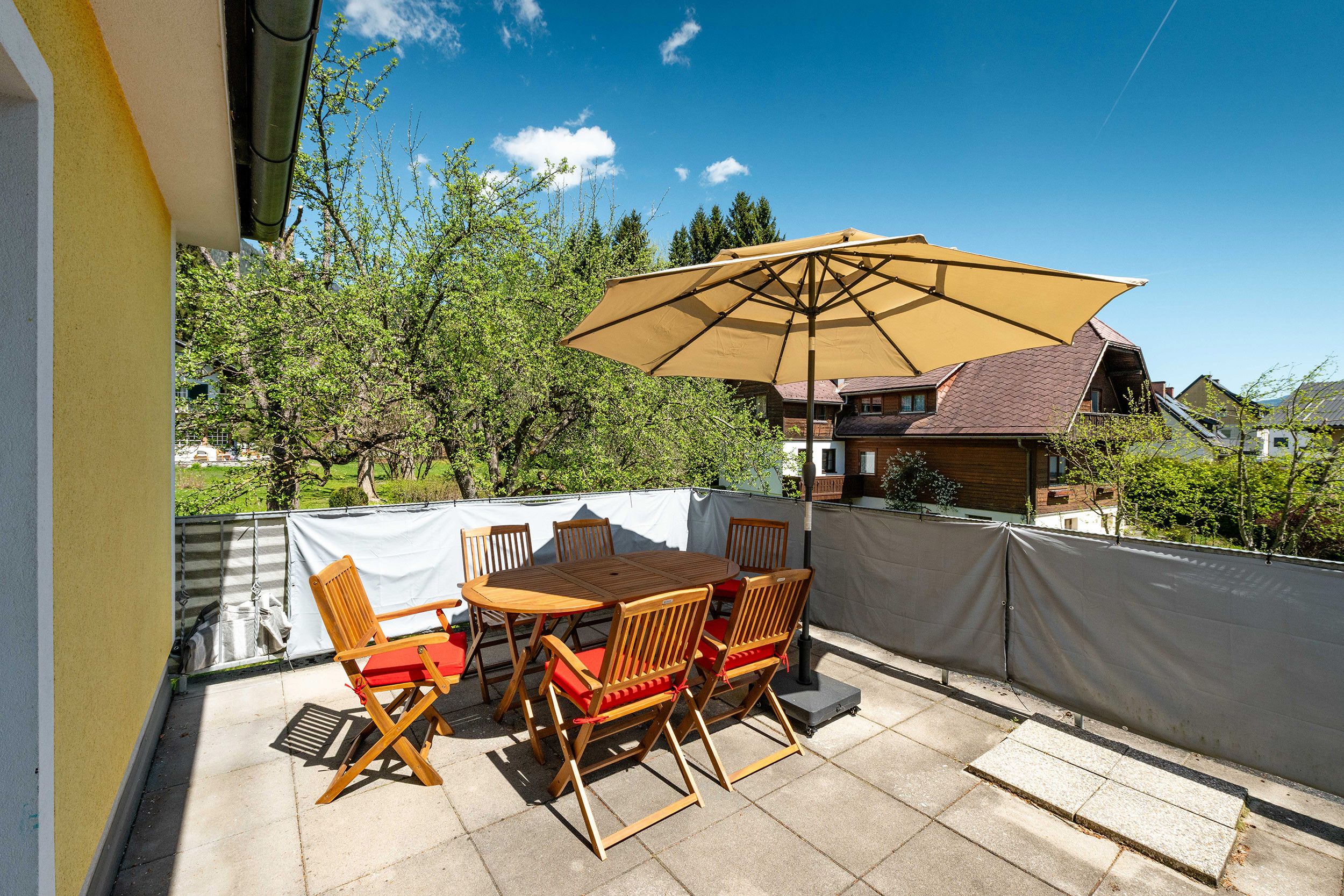An outdoor patio with a wooden table and chairs, a large umbrella, and a view of a residential area with houses and trees.