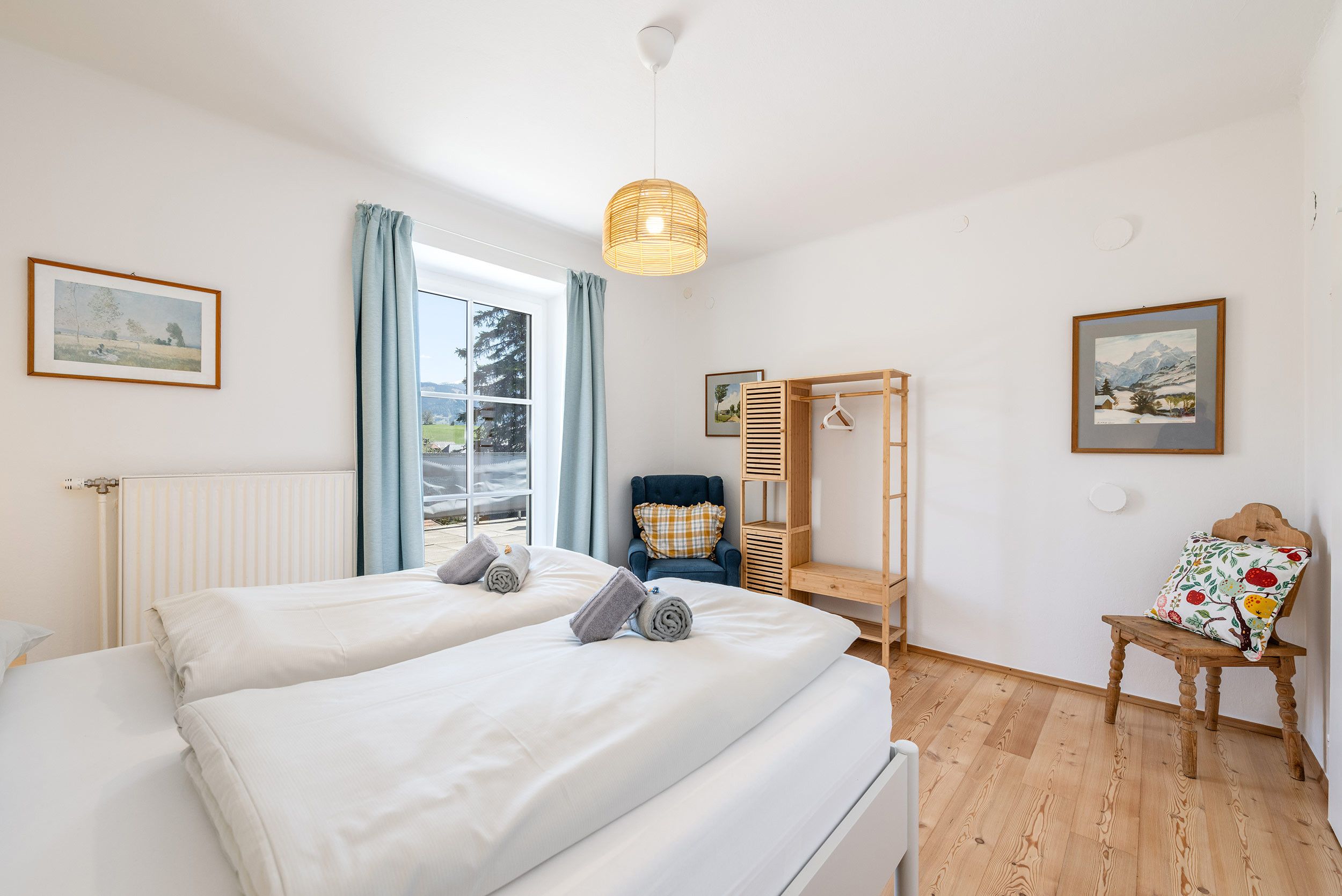 A hotel room with two beds, a wooden chair, a hanging light fixture, and a view of the outdoors.