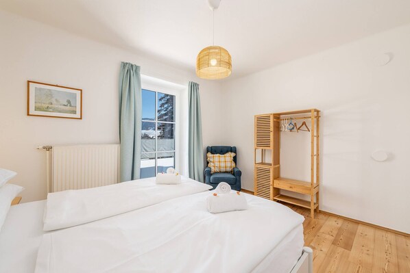 Hotelkamer met een bed, een stoel, een houten garderobe, een radiator, een raam met gordijnen en een hanglamp.