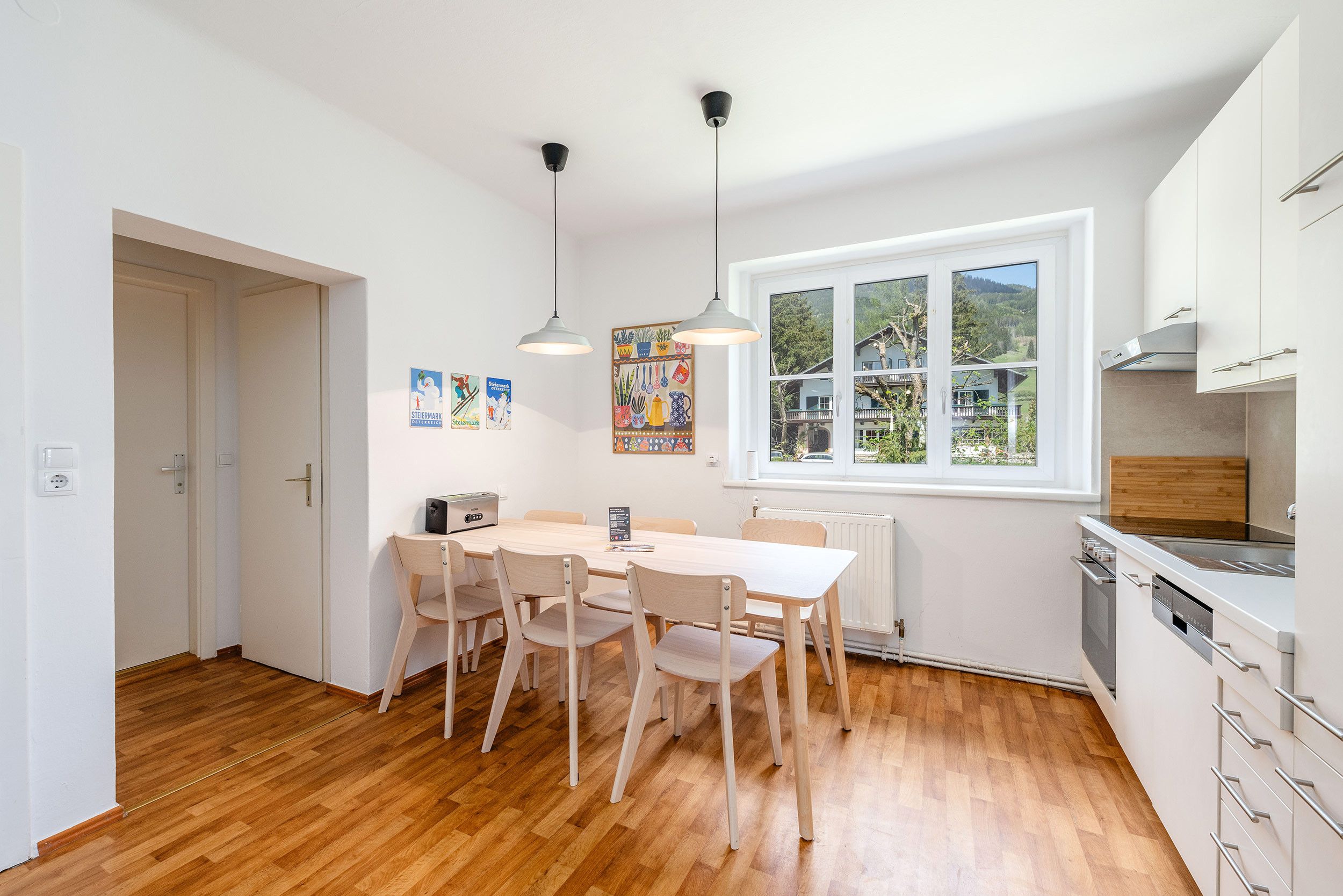 A modern kitchen with white cabinets, a wooden dining table with chairs, a built-in oven, and a window with a view of a house and trees.