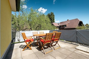 Una terrazza esterna con tavolo e sedie, una zona riparata e vista su una casa e alberi.