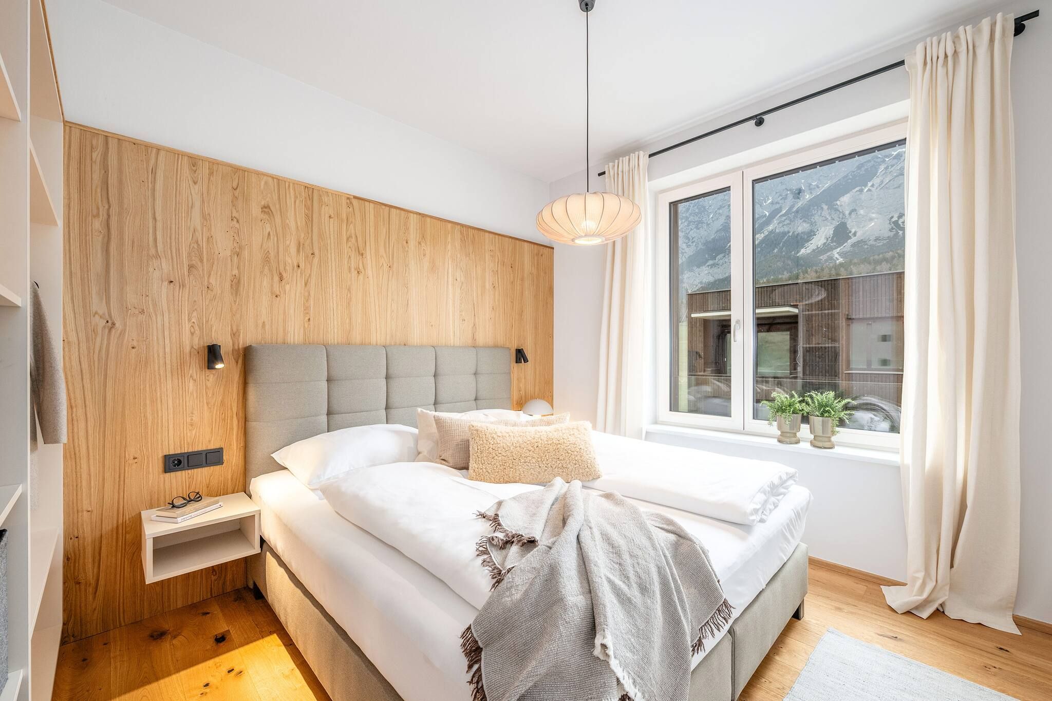 Ein modernes Schlafzimmer mit einem hölzernen Kopfteil, einem großen Bett mit weißer Bettwäsche, einem Nachttisch mit Lampe und einem Fenster mit Vorhängen.