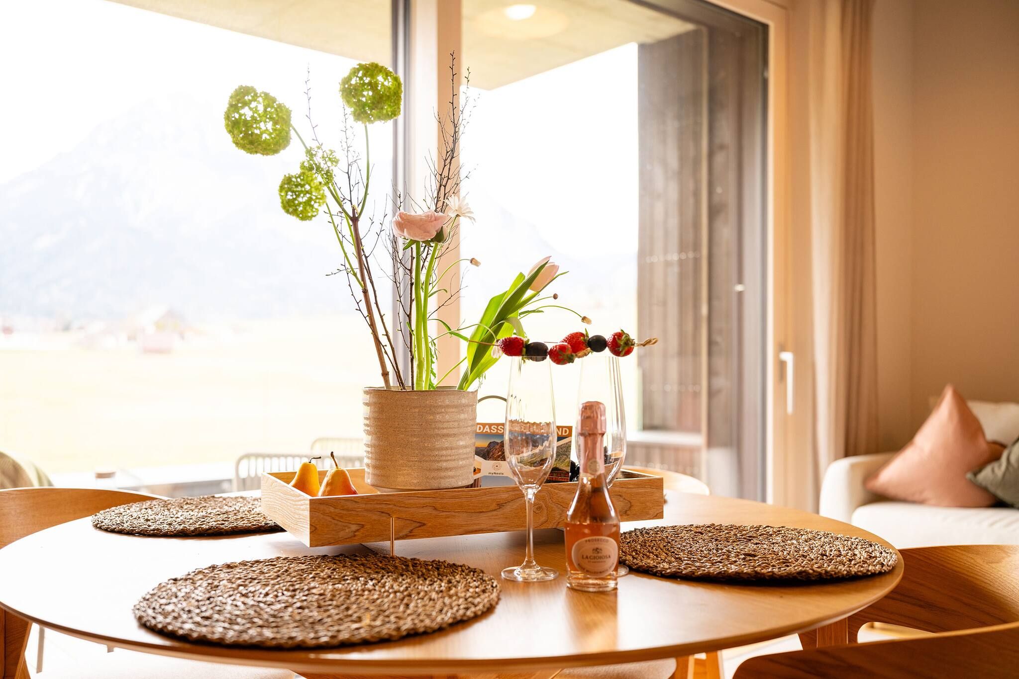Une table ronde en bois, surmontée d’un set de table en fibres tressées, un vase contenant des fleurs vertes en forme de sphères, une bouteille en verre et un livre intitulé « Mon nid ».