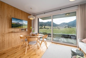 Une pièce dotée d’un plancher en bois, d’une table à manger avec des chaises, d’un canapé, d’un téléviseur fixé au mur et d’une grande fenêtre donnant sur un paysage verdoyant.