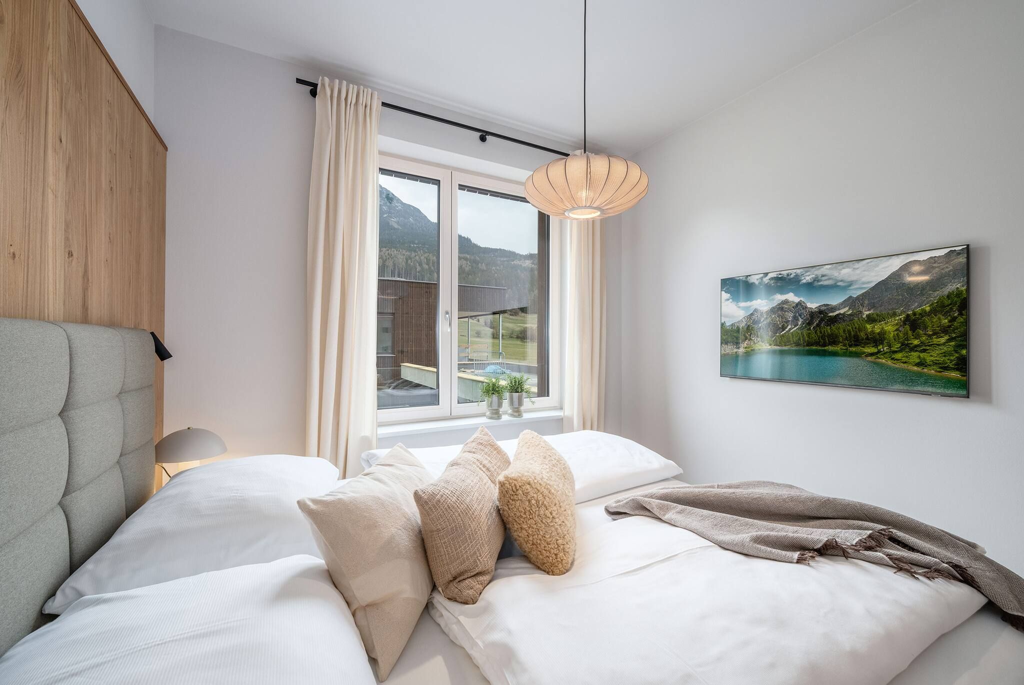 Ett sovrum med en säng, kuddar, ett fönster med gardiner, en hängande lampa och en landskapsmålning på väggen.