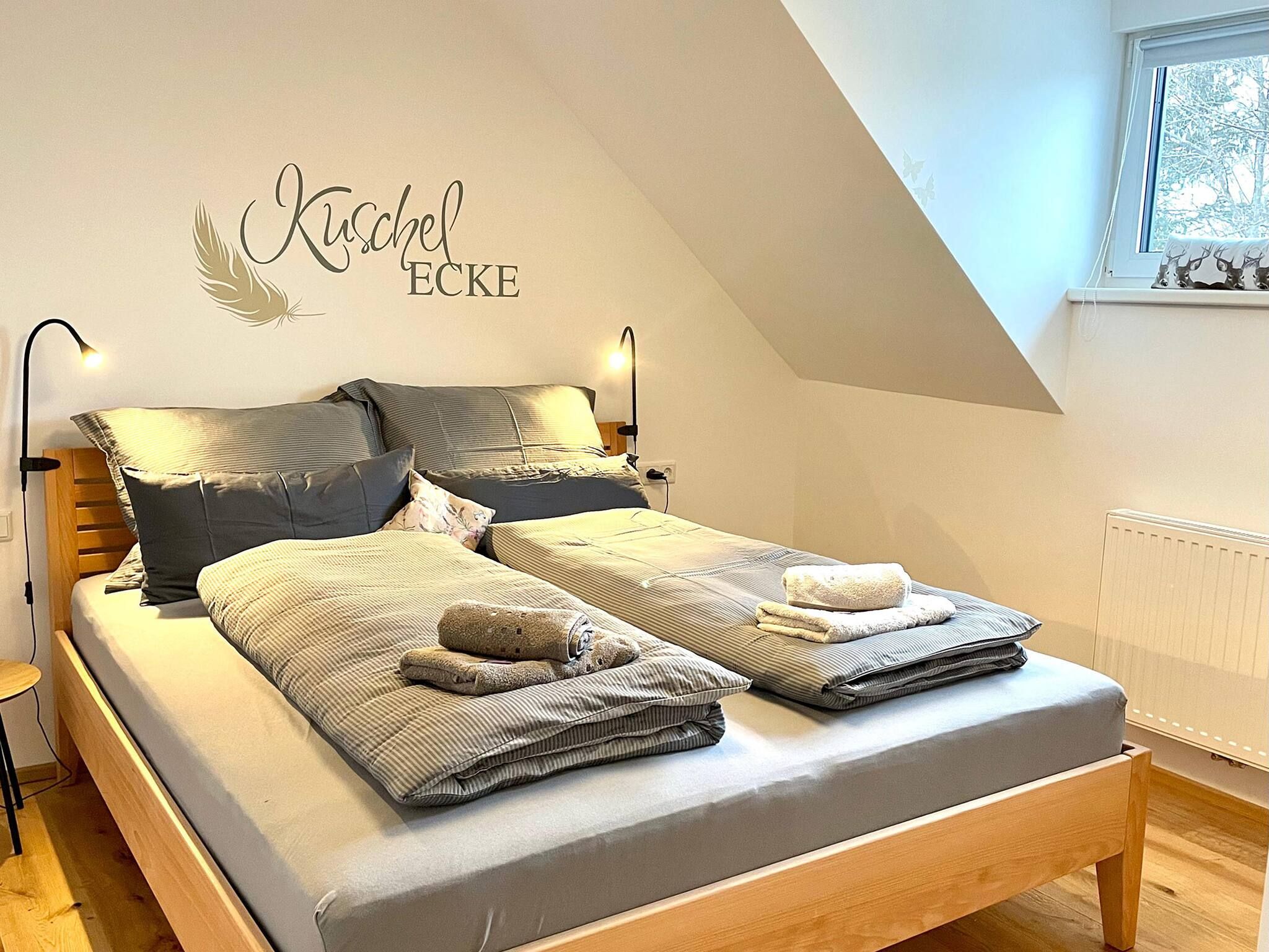 Une chambre avec un plafond en pente, un lit agrémenté de plusieurs oreillers et de couvertures, une lampe fixée au mur et une fenêtre donnant sur des arbres.