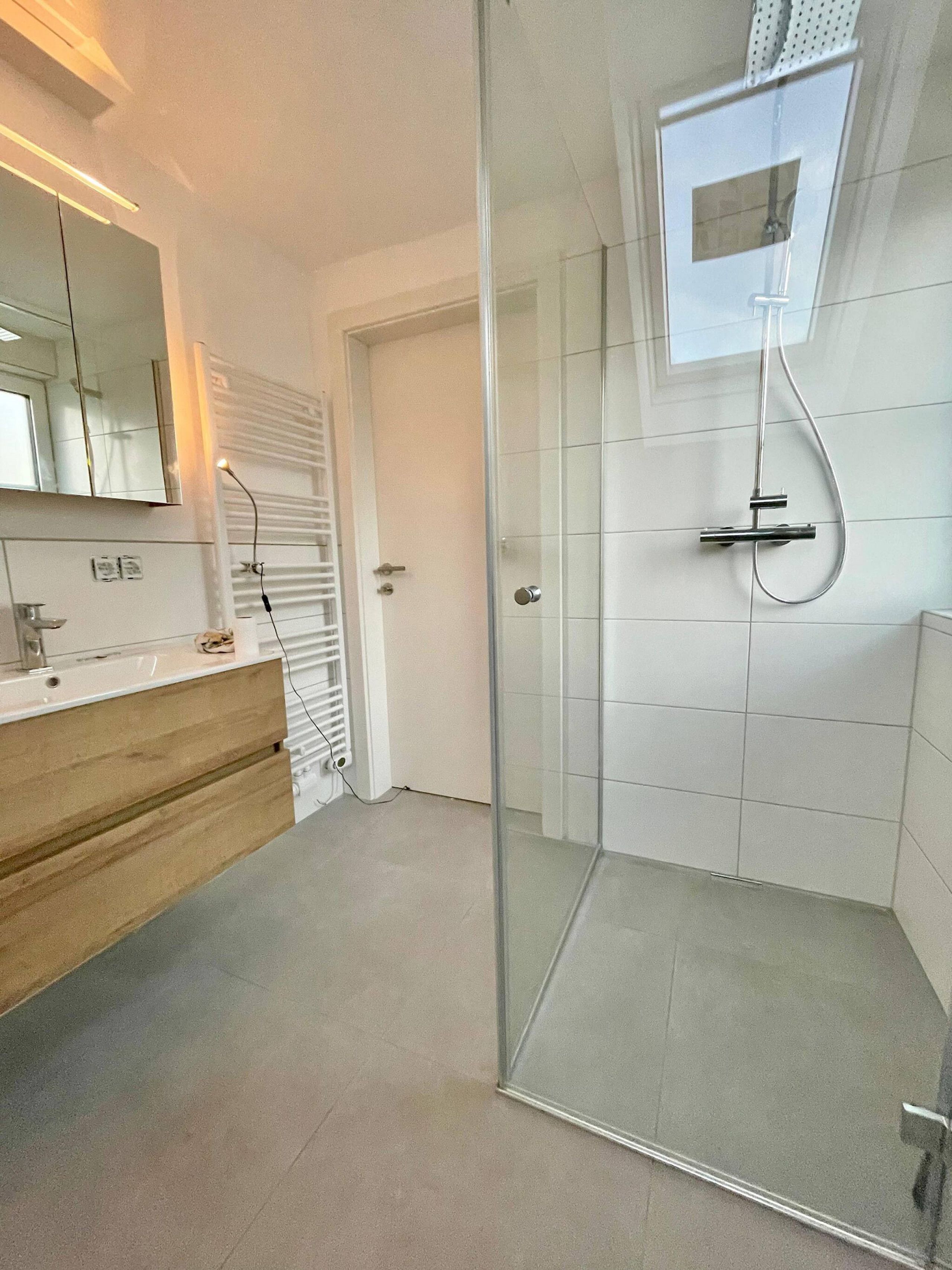 Ein modernes Badezimmer mit Duschkabine aus Glas, weiß gefliester Wand und Waschtisch mit Spiegel.