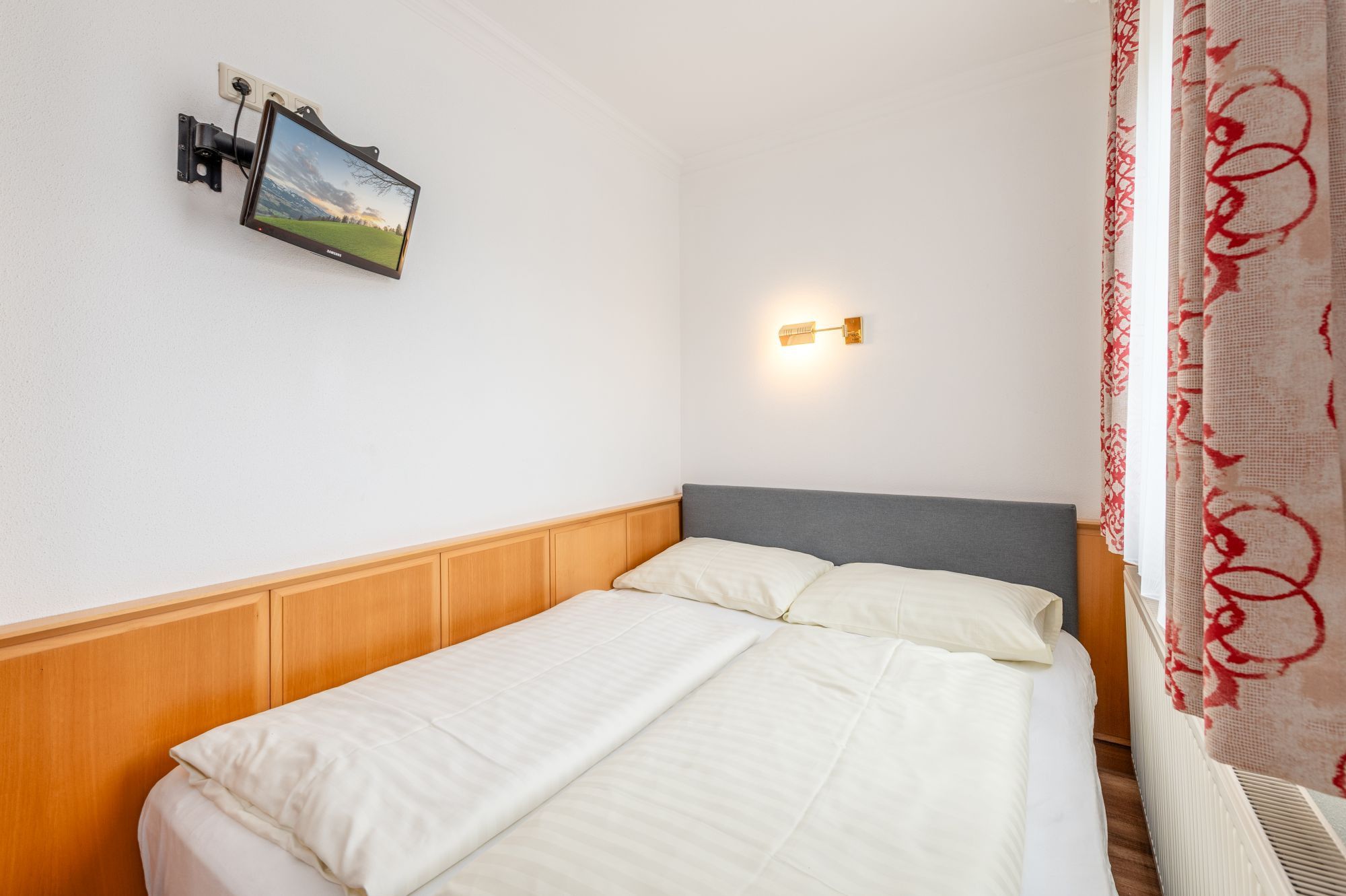 Une chambre d’hôtel avec un lit simple doté d’une tête de lit en bois, un téléviseur fixé au mur et une lampe de chevet.