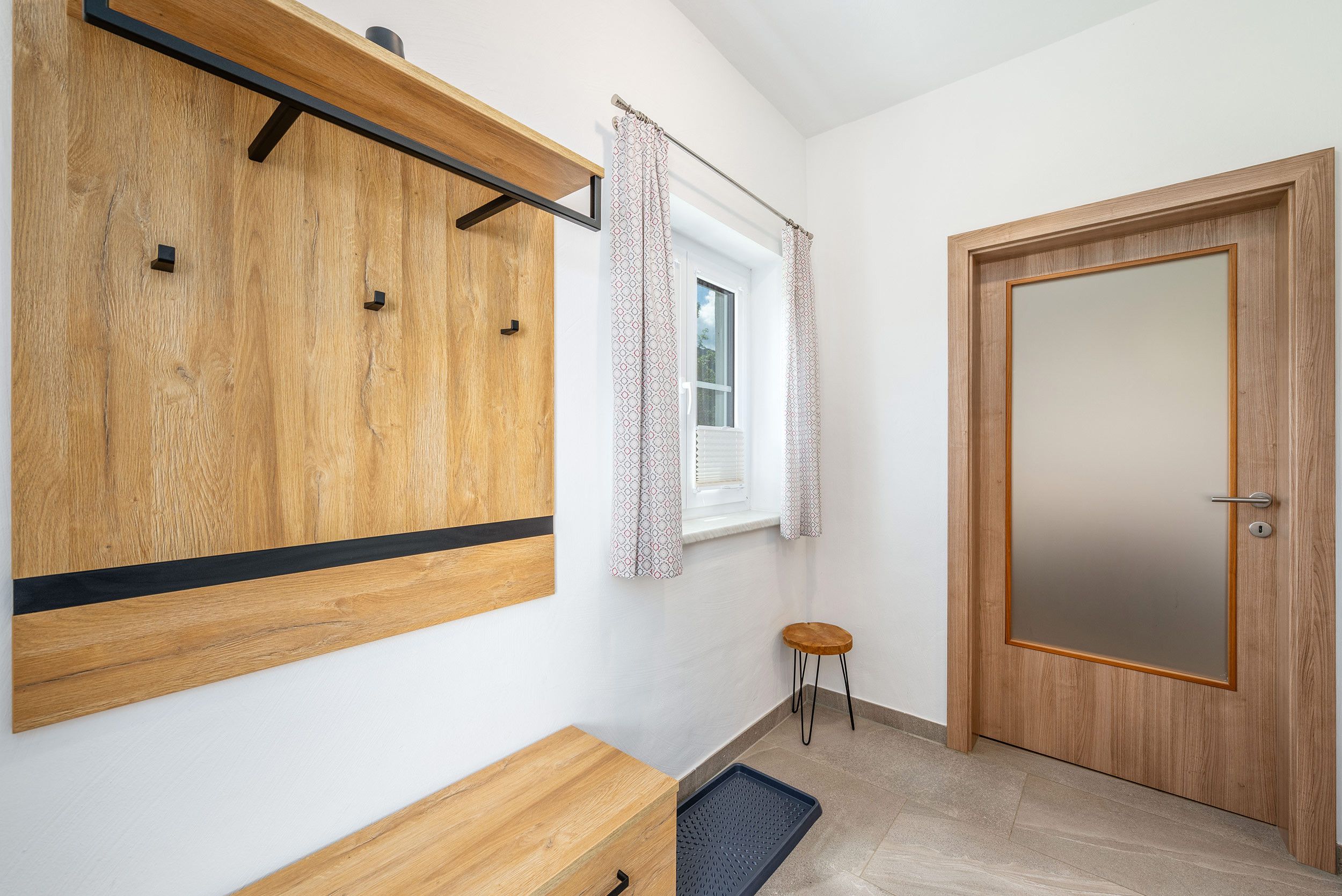 Ein Zimmer mit einem Holzschrank, einem kleinen Hocker, einer Tür und einem Fenster mit gemusterten Vorhängen.