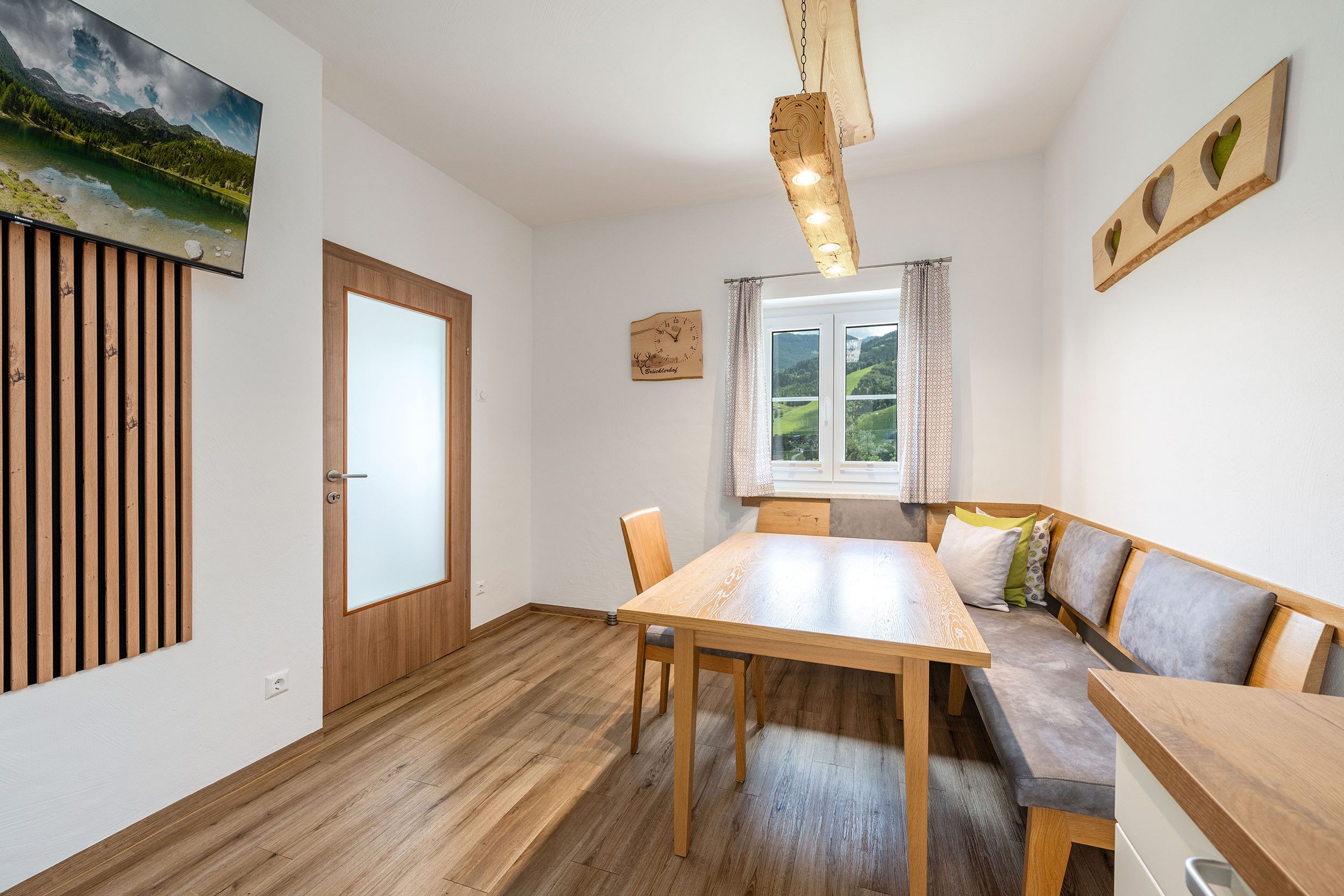 Ein Zimmer mit einem Holztisch, einer Bank, einem Fenster mit Blick auf Grünflächen und einer Holzverkleidung an der Wand.