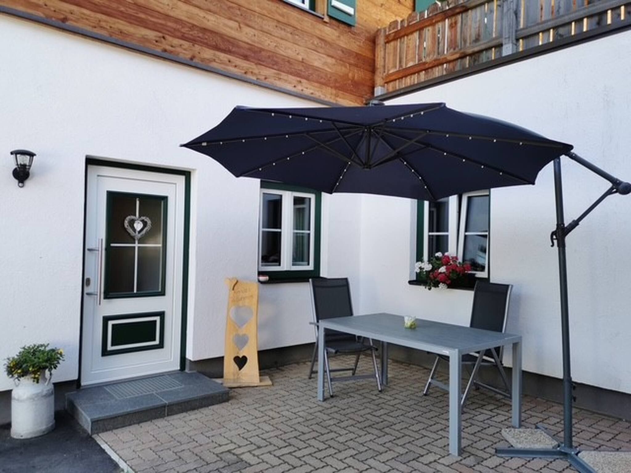 Un espace patio avec une table et des chaises, un grand parasol, une porte décorée d’un motif en forme de cœur et une jardinière.