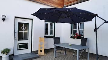 Un espace patio avec une table et des chaises, un grand parasol, une porte décorée d’un motif en forme de cœur et une jardinière.