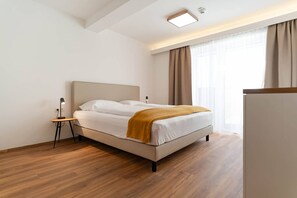 Ein modernes Hotelzimmer mit Bett, Nachttisch, Lampe und Blick ins Freie durch ein Fenster.