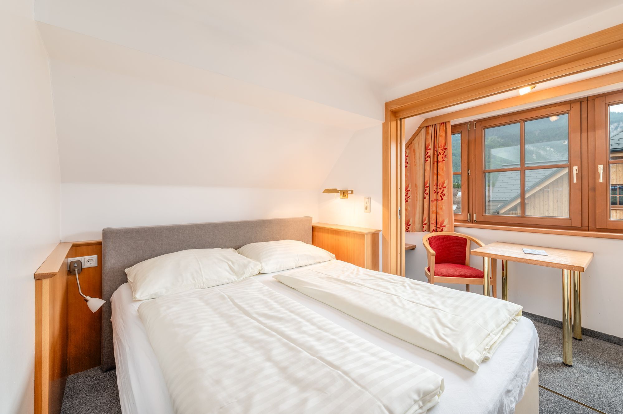 Una camera d'albergo con un letto, un comodino, una lampada, una sedia e una finestra con tende.