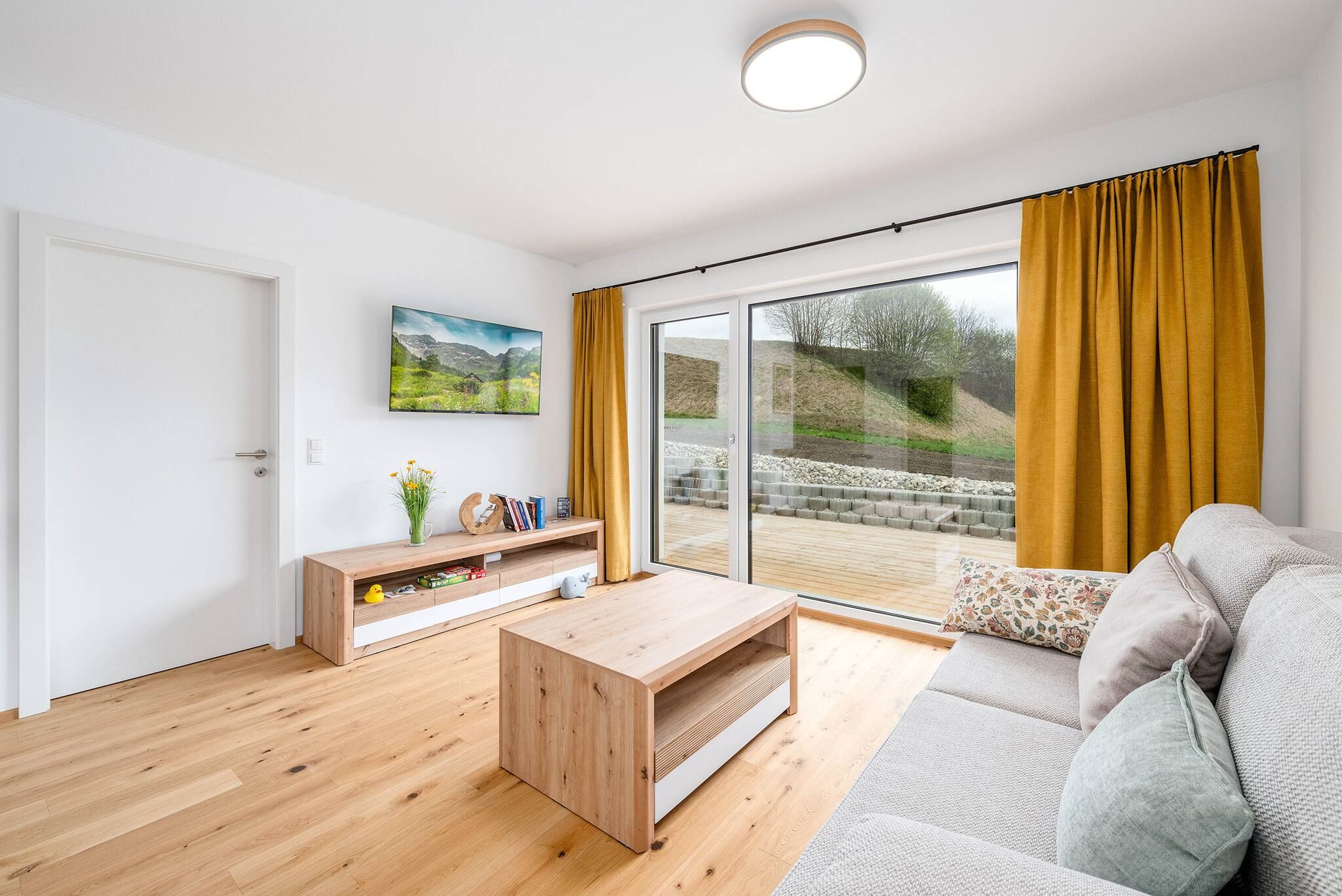 Ein modernes Wohnzimmer mit Holzboden, einer Couch, einem Couchtisch, einem Fernseher und einer Glastür zum Balkon.