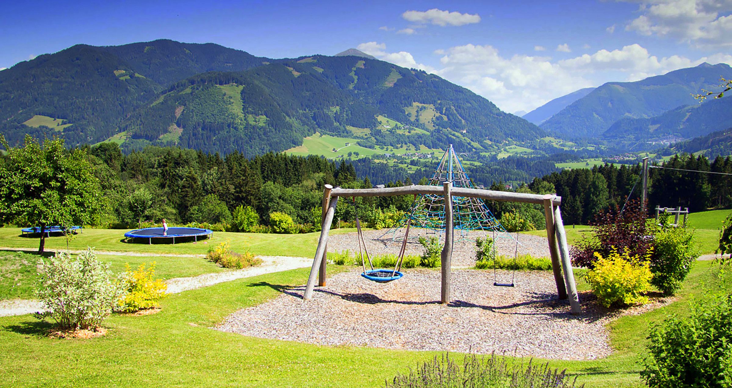 A playground with swings and a trampoline, surrounded by a grassy area and trees, with mountains in the background.