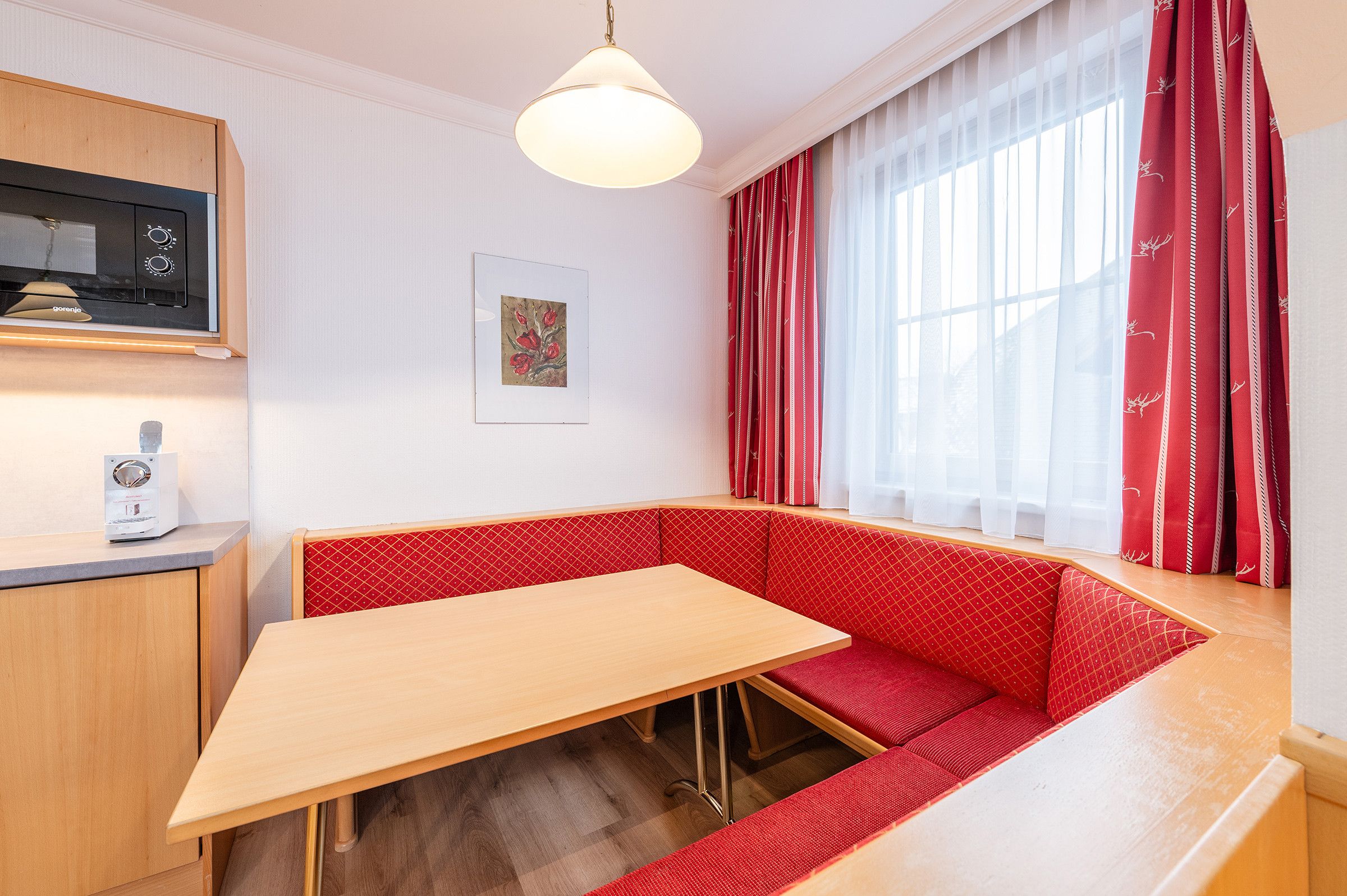 Una stanza con tavolo in legno, sedute su una panca imbottita di rosso, un forno a microonde e una finestra con tende rosse.