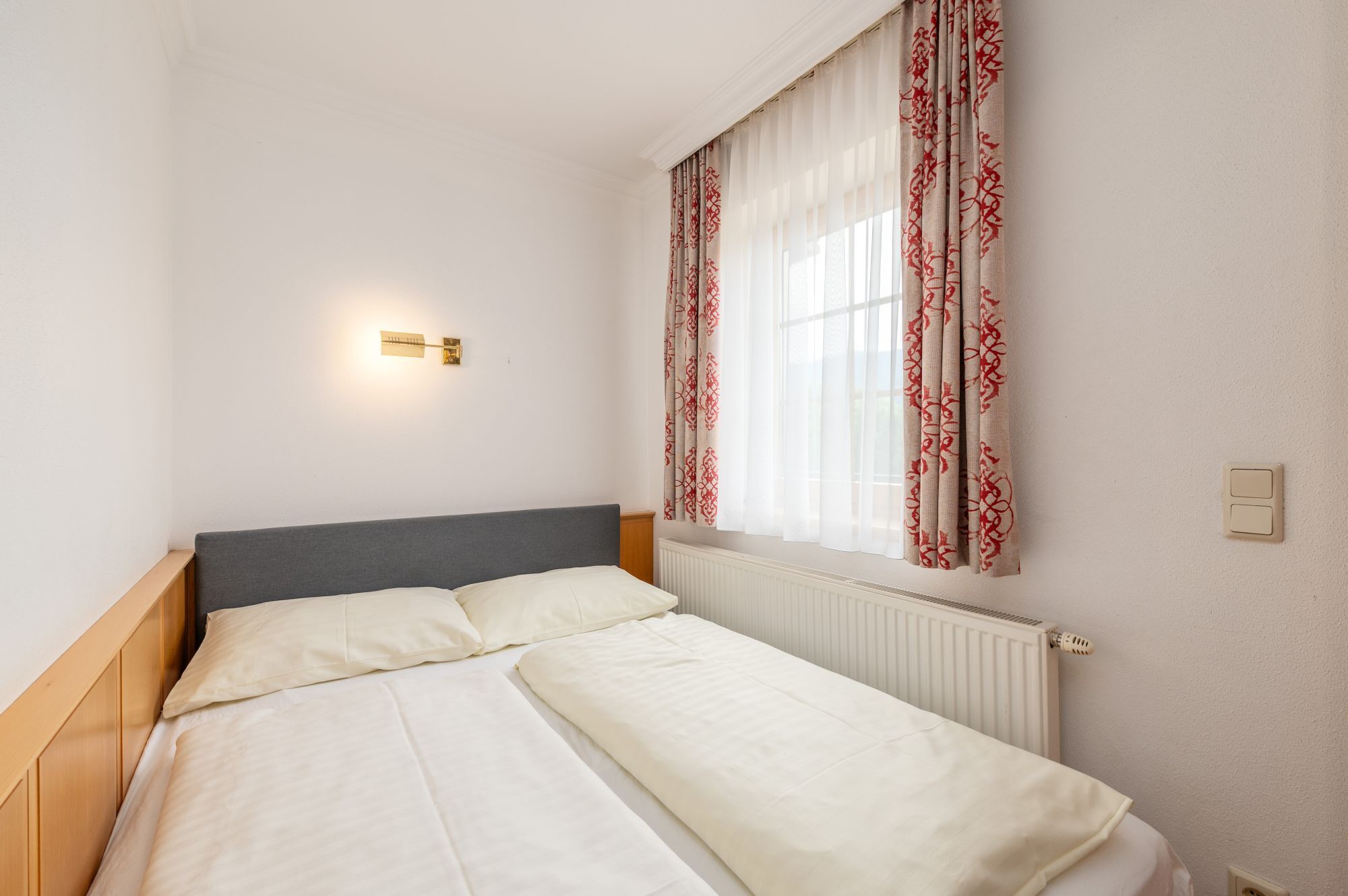 Ein Einzelbett mit grauem Kopfteil, weißer Bettwäsche und einem Fenster mit roten, gemusterten Vorhängen.