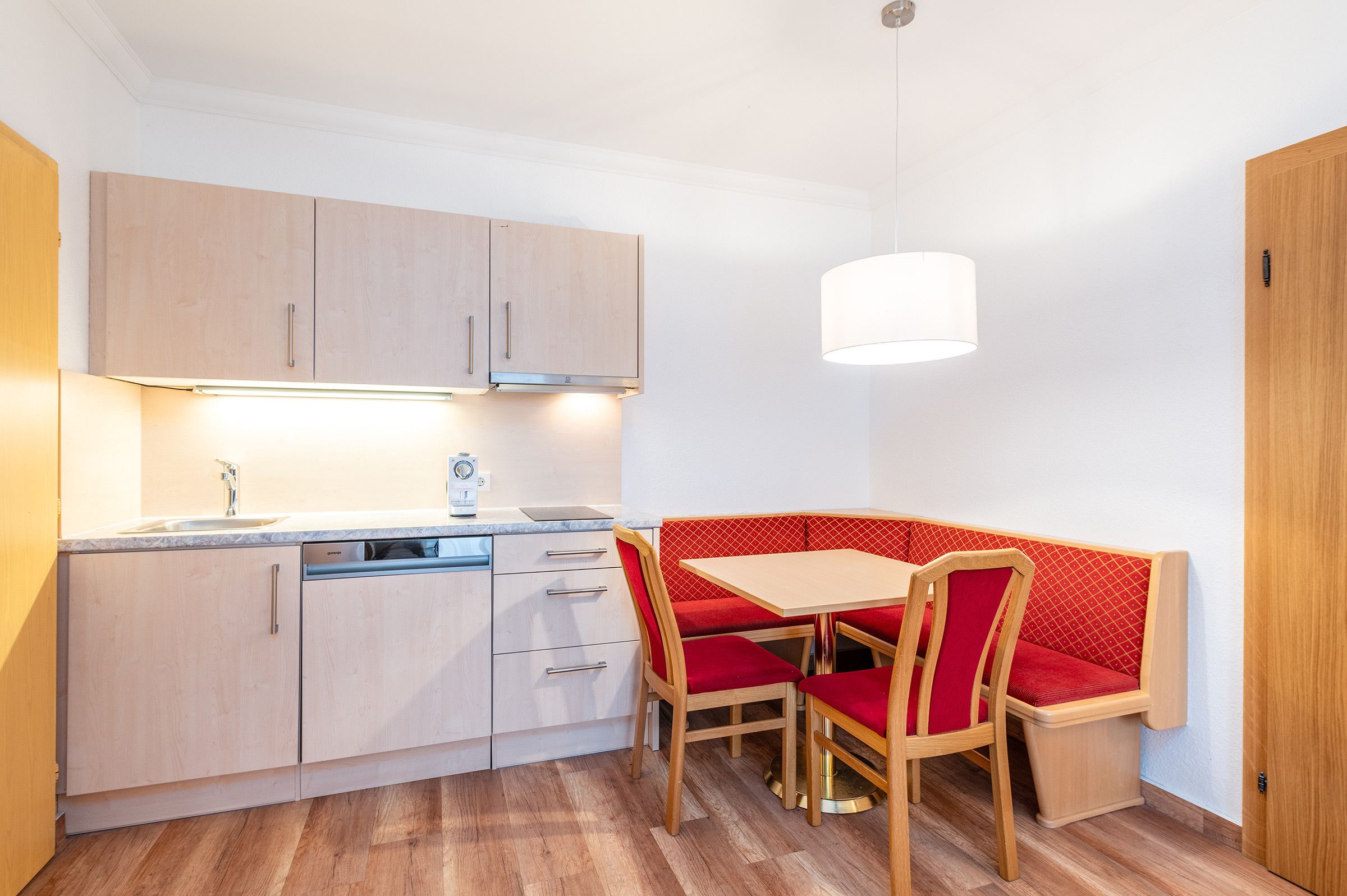 Cocina compacta con zona de comedor, que incluye lavavajillas integrado, armarios de madera y una banca tapizada en rojo.