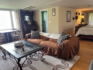 Un salon chaleureux avec un canapé marron, une table basse en verre, un piano et un lit à l’arrière-plan.