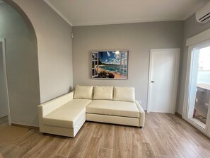 Habitació amb un sofà seccional beix, un quadre amb una escena de platja, una porta i una finestra.