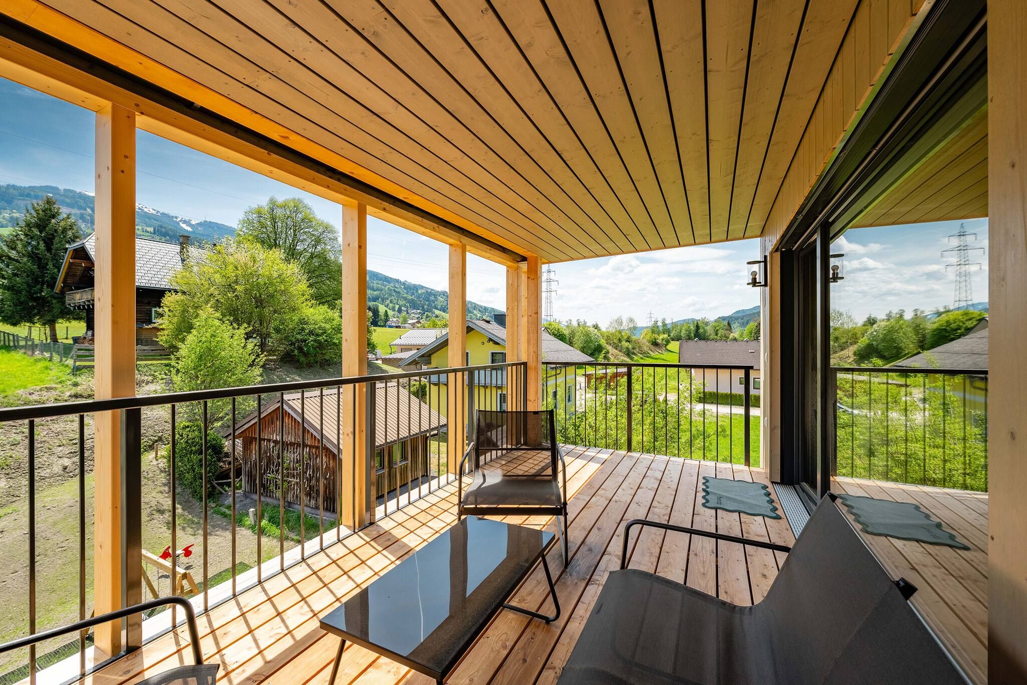 Eine Holzterrasse mit Glasgeländer, einem Metallstuhl und Blick auf eine ländliche Landschaft mit Häusern und Bäumen.