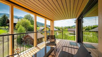 Eine Holzterrasse mit Glasgeländer, einem Metallstuhl und Blick auf eine ländliche Landschaft mit Häusern und Bäumen.