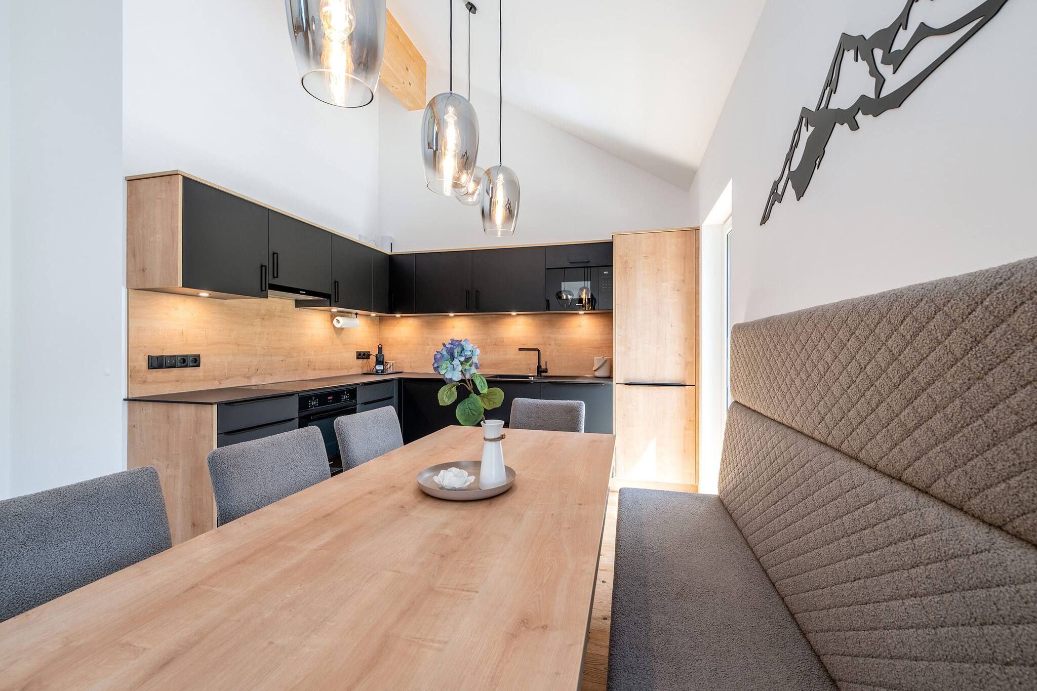 Une cuisine moderne avec une table à manger en bois, des chaises à revêtement gris et des luminaires suspendus.