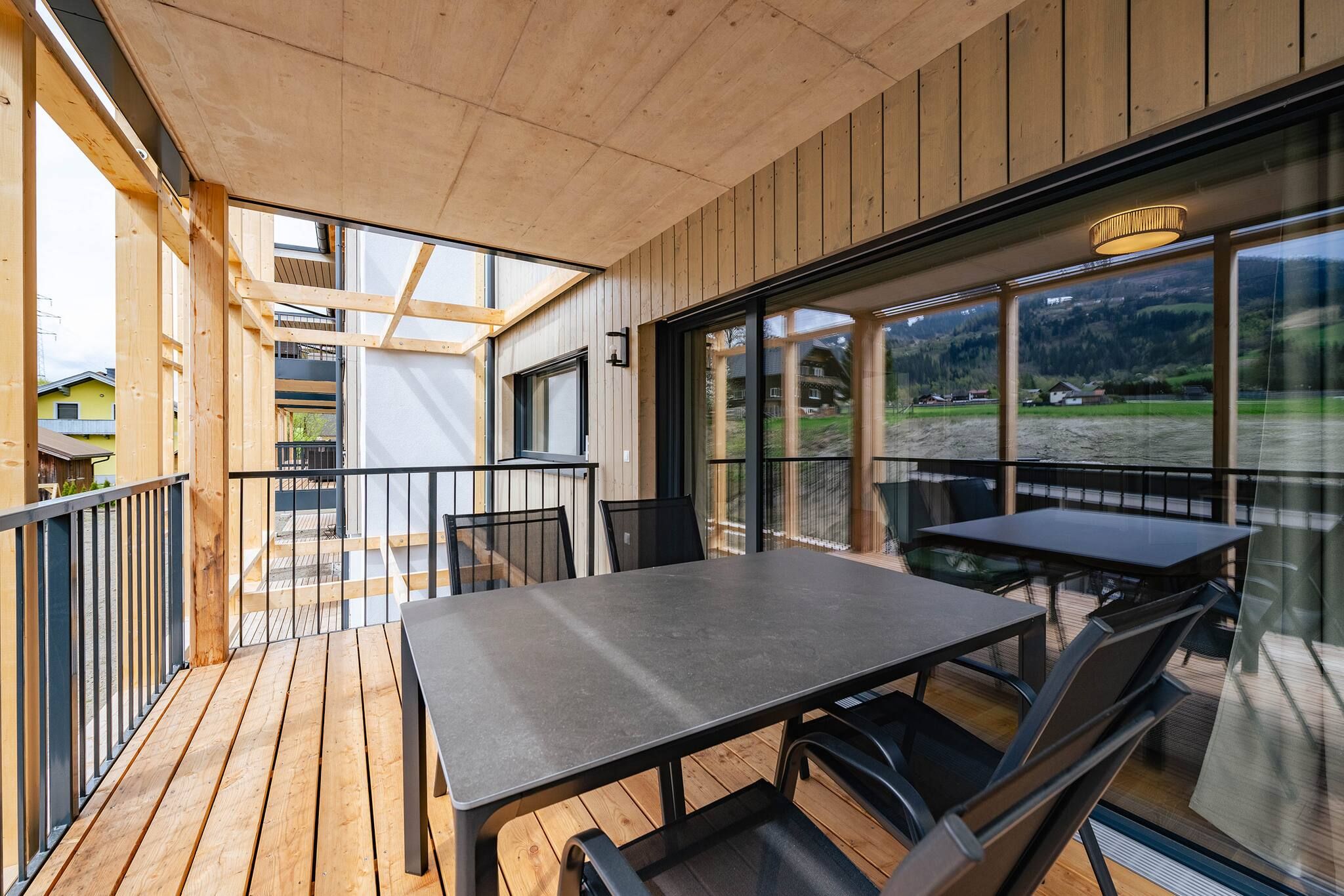 Eine Holzterrasse mit Tisch und Stühlen, große Fenster mit Blick auf eine gebirgige Landschaft und ein Balkon mit Geländer.