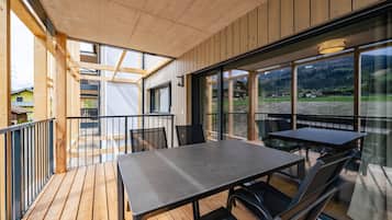 Eine Holzterrasse mit Tisch und Stühlen, große Fenster mit Blick auf eine gebirgige Landschaft und ein Balkon mit Geländer.