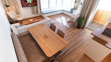 A modern living room with a wooden dining table, cushioned seating, a flat-screen TV, and a large window with curtains.