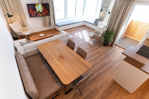 Ein modernes Wohnzimmer mit einem Holzesstisch, bequemen Sitzgelegenheiten, einem Flachbildfernseher und einem großen Fenster mit Vorhängen.