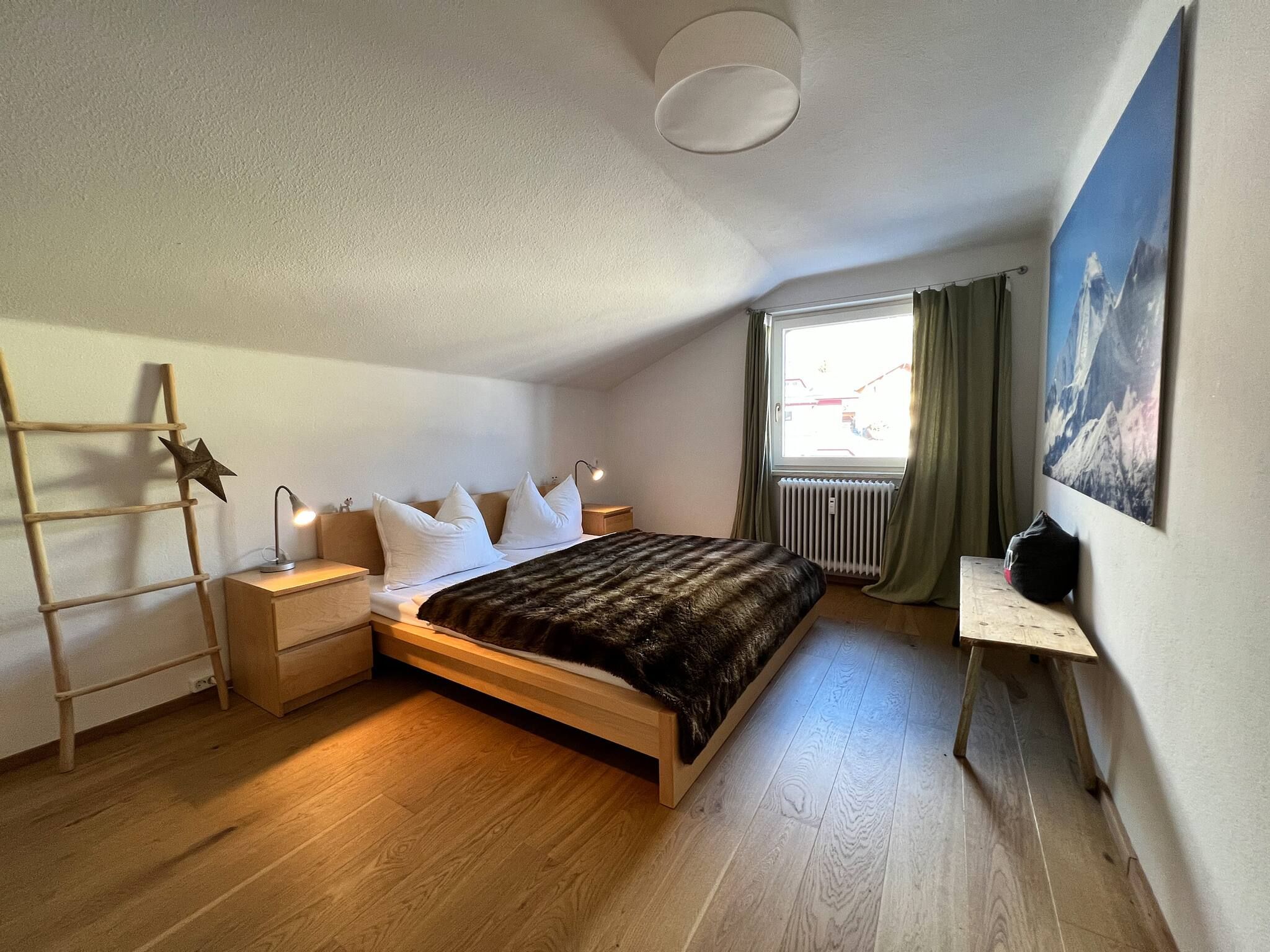 Ein Schlafzimmer mit einem hölzernen Bett, einem Nachttisch, einer Lampe, einer Leiter, einer Heizung, einem Fenster mit Vorhängen und einem Bild an der Wand.