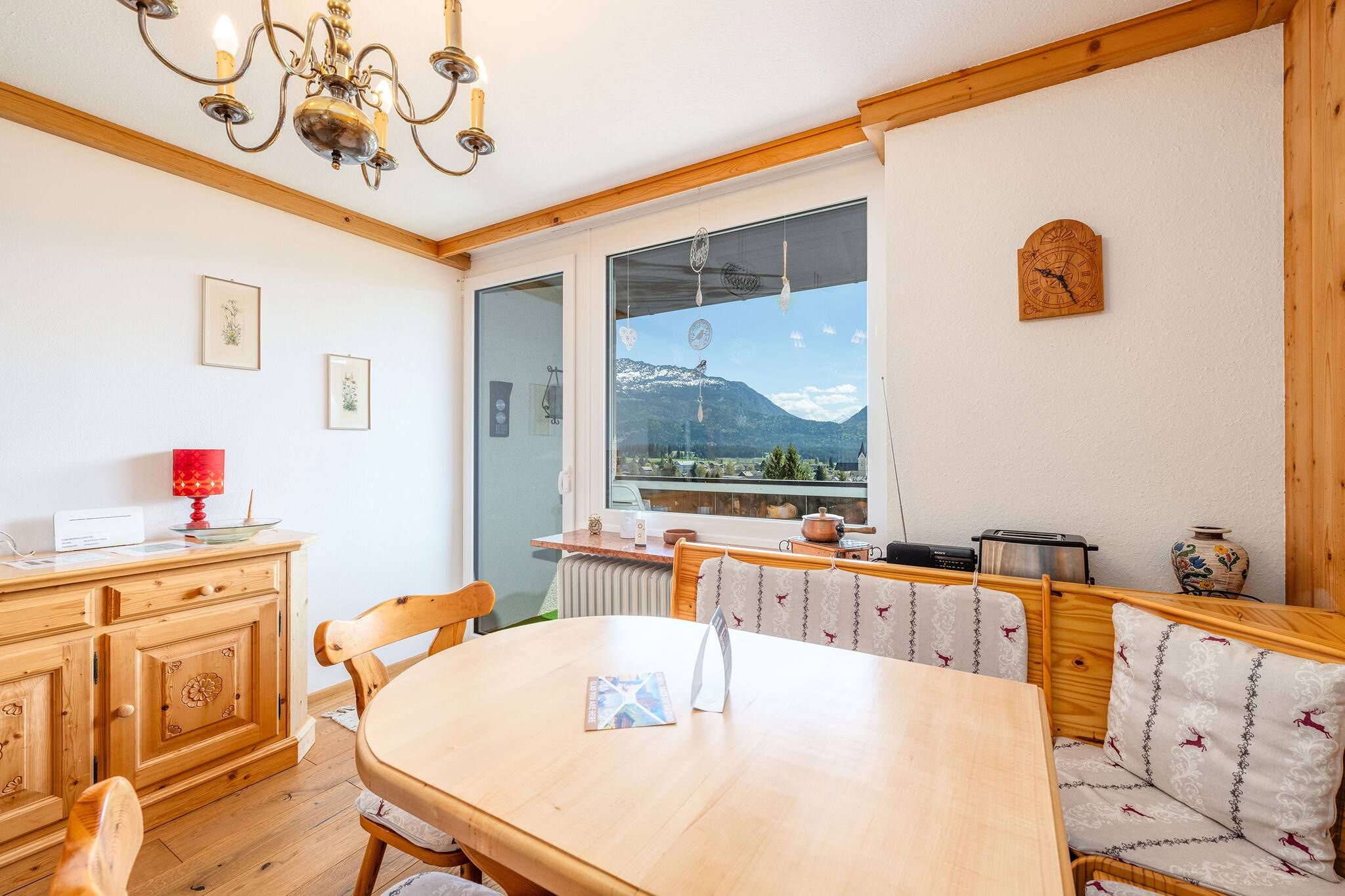 A cozy dining area with a wooden table and chairs, a chandelier, and a view of mountains through a large window.