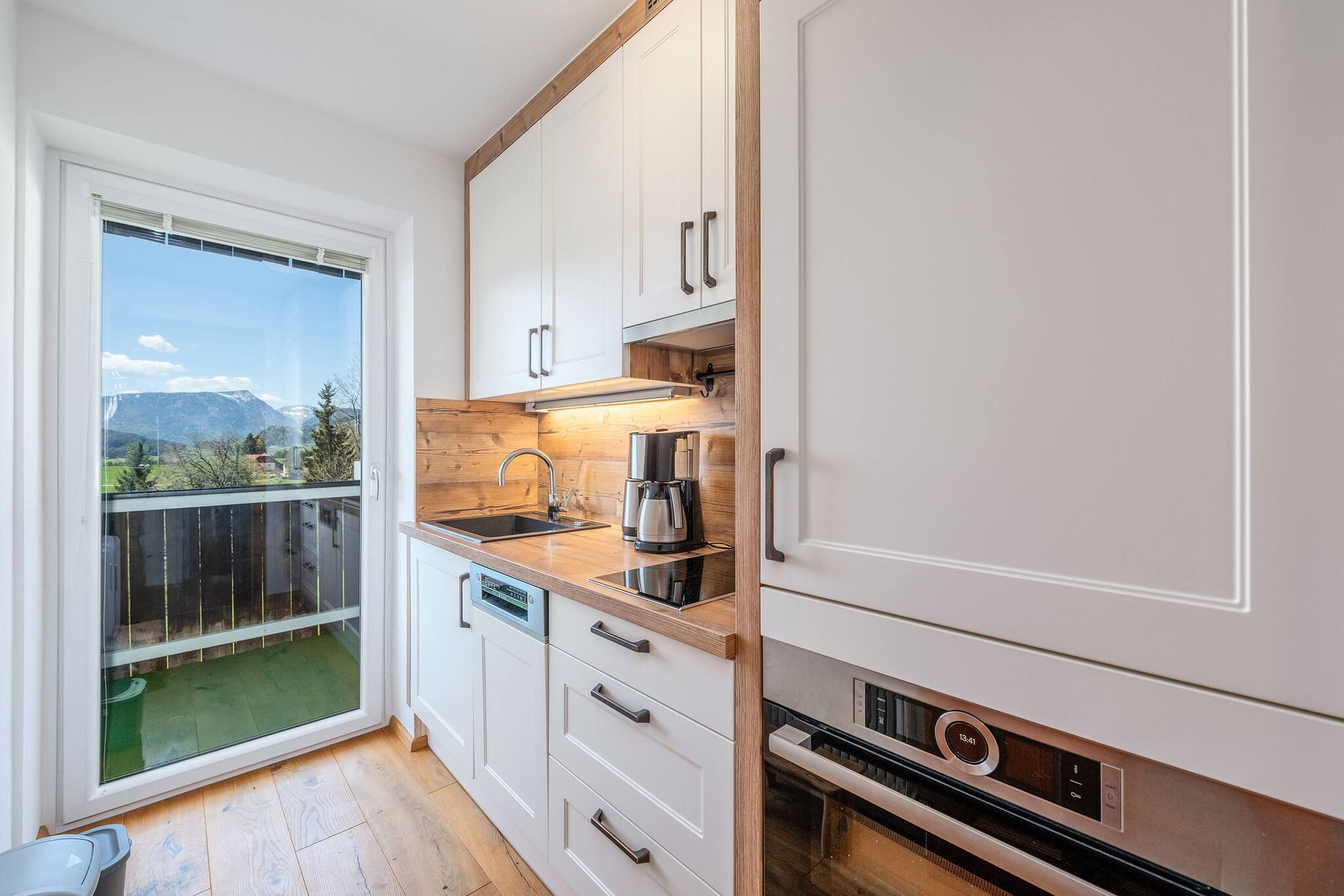 A compact kitchen with white cabinets, a wooden countertop, a built-in oven, and a view of a mountainous landscape through a sliding glass door.