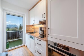 A compact kitchen with white cabinets, a wooden countertop, a built-in oven, and a view of a mountainous landscape through a sliding glass door. - Alpenblick Bad Mitterndorf 30 (Bad Mitterndorf)