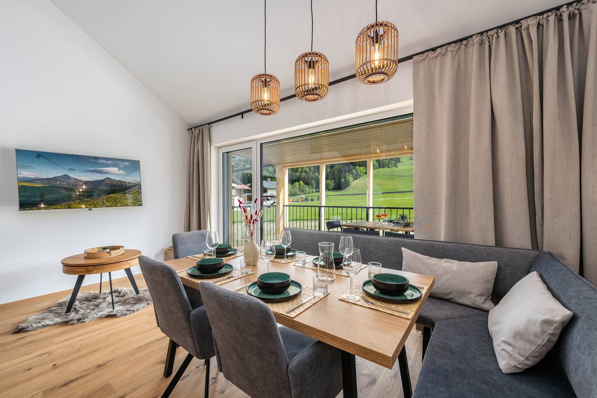 Un coin repas avec une table dressée pour quatre personnes, un canapé et une vue sur un paysage verdoyant grâce à de grandes fenêtres.