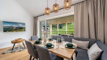Un coin repas avec une table dressée pour quatre personnes, un canapé et une vue sur un paysage verdoyant grâce à de grandes fenêtres.