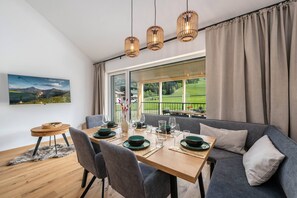 Ein Essbereich mit einem Tisch für vier Personen, einer Couch und Blick auf eine grüne Landschaft durch große Fenster.