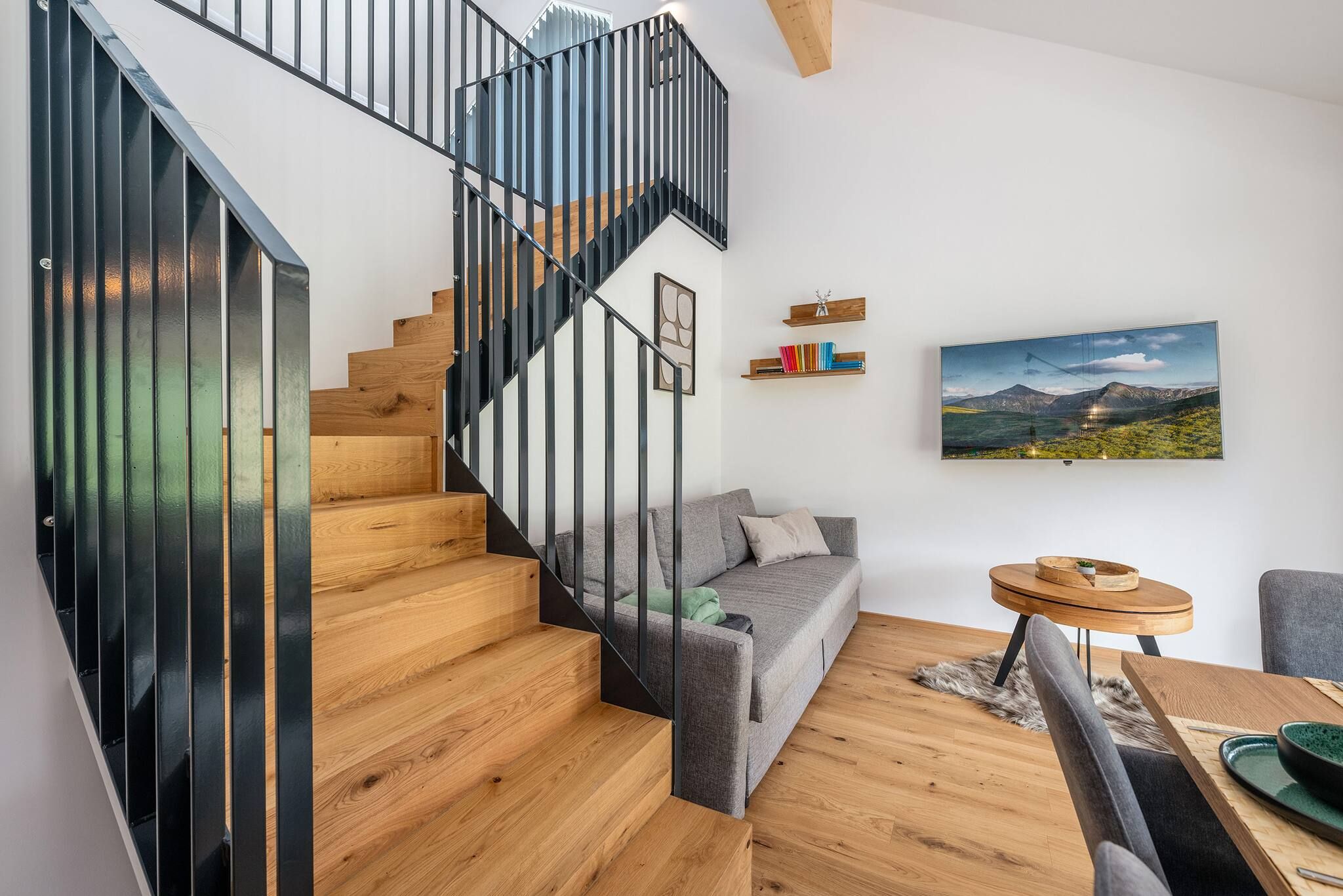 Ein modernes Interieur mit einer Holztreppe, einem grauen Sofa, einem Holztisch und einem Gemälde einer Berglandschaft an der Wand.