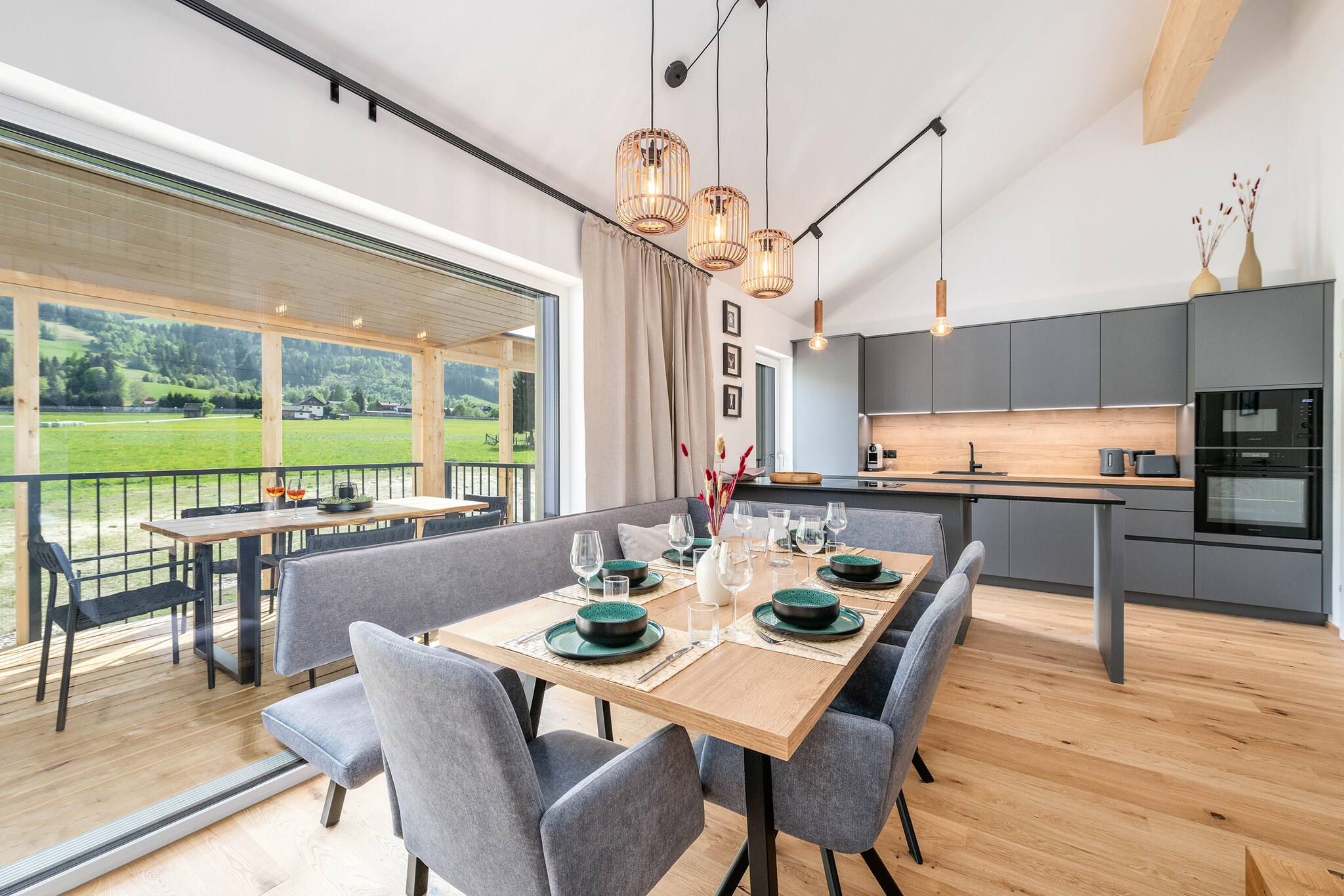 Un espace repas moderne avec une table en bois dressée pour six personnes, des chaises à revêtement gris et une vue sur un paysage verdoyant grâce à de grandes fenêtres.