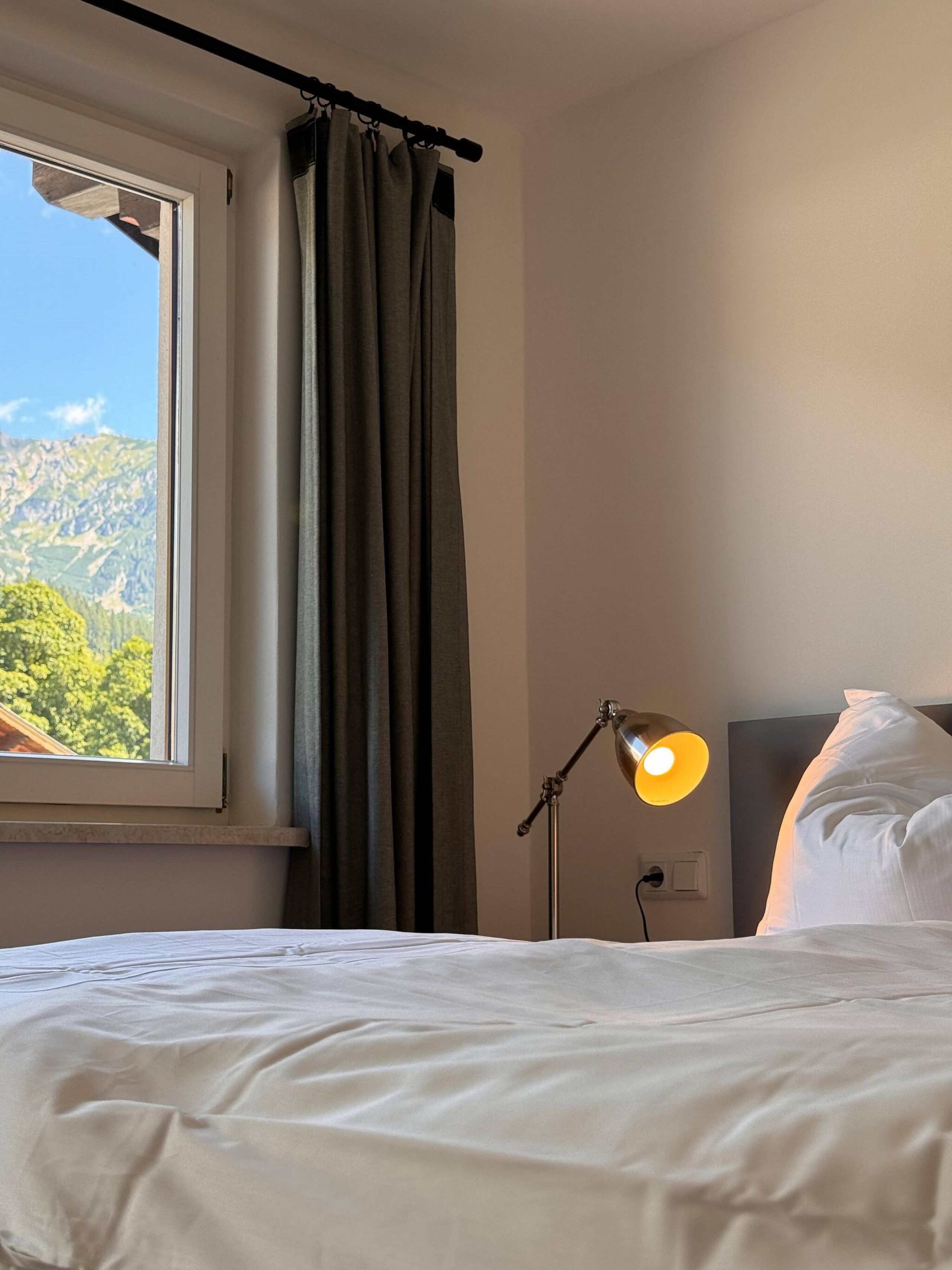 Ein Hotelzimmer mit einem Bett, einem Fenster mit Vorhängen, einer Lampe und Bergblick.
