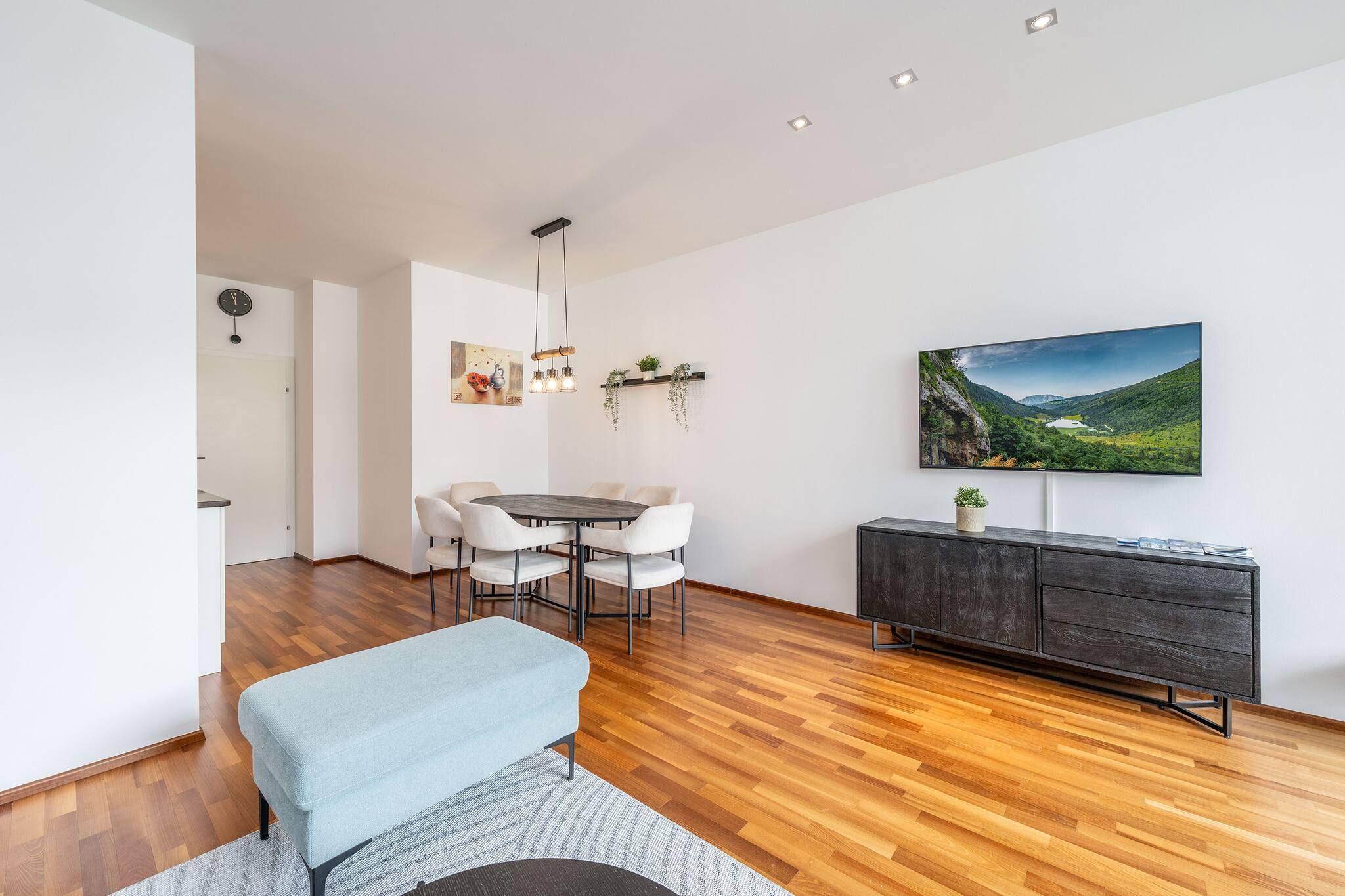 Uma sala de estar moderna com área de jantar, piso de madeira, um sofá cinza, uma TV na parede e um armário preto.